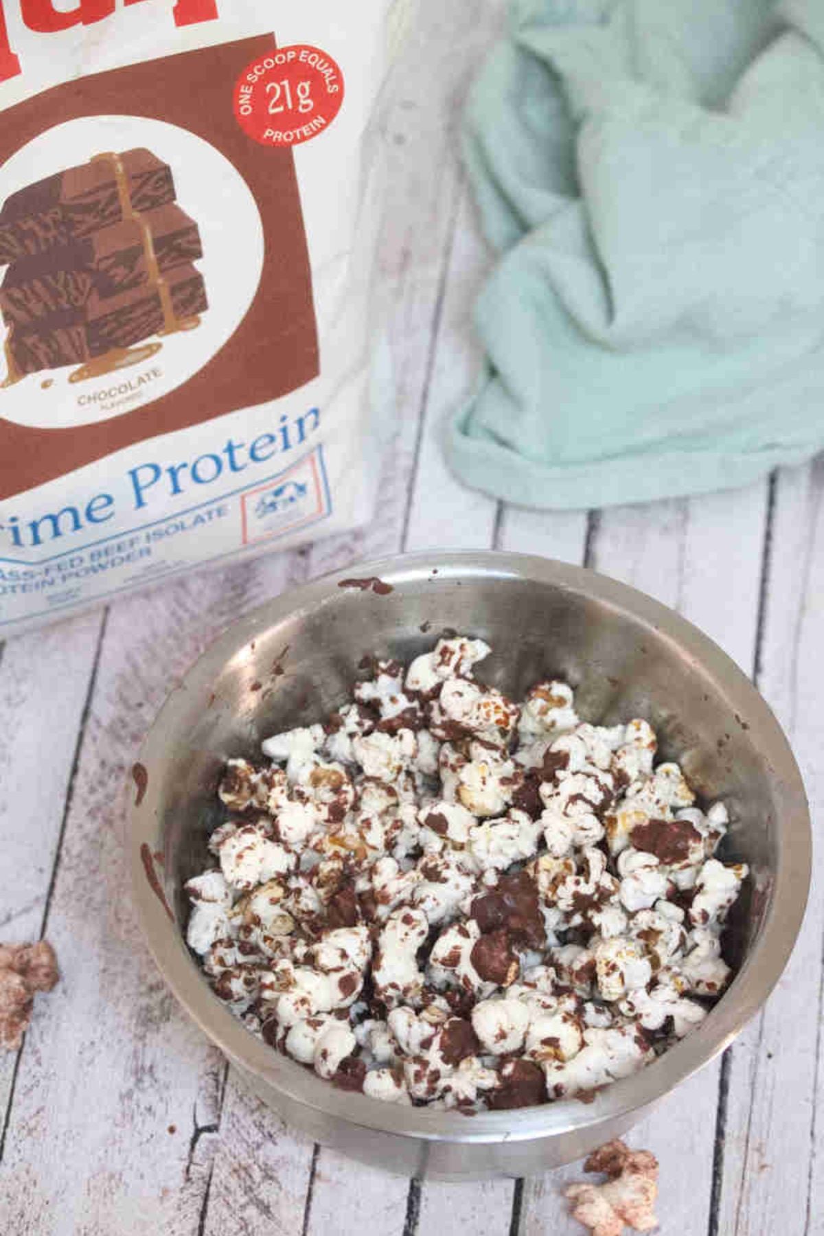 Steel bowl with chocolate drizzled protein popcorn in it and a bag of protein behind it.