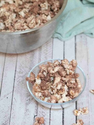 Chocolate protein powder popcorn in a glass bowl with more popcorn in a large bowl behind it.