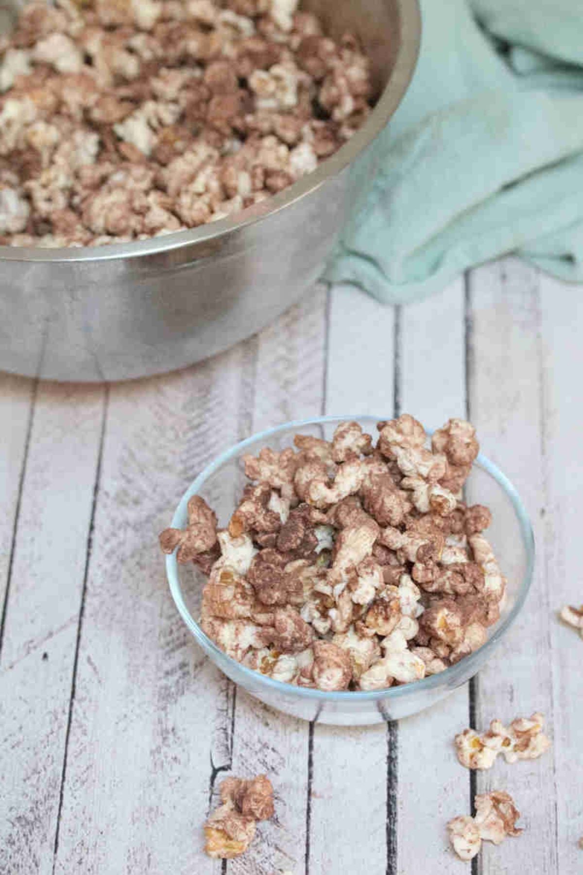 Chocolate protein powder popcorn in a glass bowl with more popcorn in a large bowl behind it.