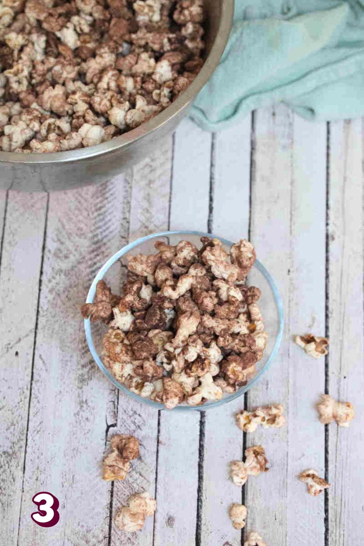 Chocolate protein powder popcorn in a glass bowl with more popcorn in a large bowl behind it.